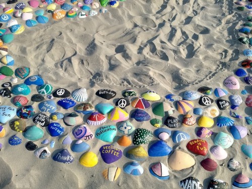 Colorfully painted rocks arranged in a sandy area, featuring various messages and designs.