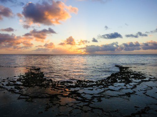 Sunset over the ocean, with colorful clouds reflecting on the water and rocky shoreline in the foreground.