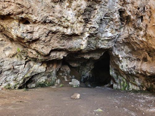 A rocky cave entrance with a sandy floor and moss-covered walls, surrounded by natural stone formations.