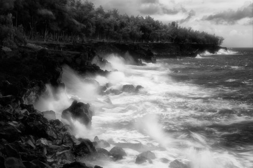 Black and white image of waves crashing against rocky coastline under a cloudy sky.