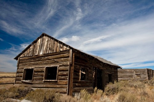 Abandoned wooden building in a desolate landscape under a blue sky with wispy clouds.