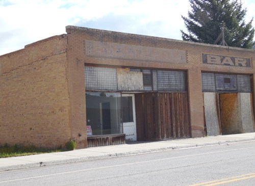 Abandoned brick building with boarded windows, featuring a faded sign that reads "BAR." Overgrown grass in front.