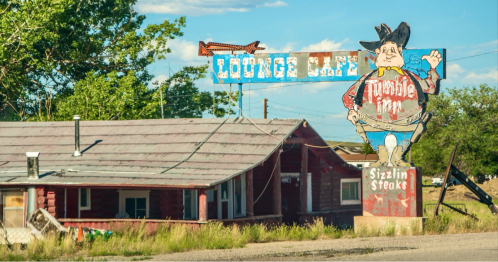 A rustic cafe with a large cowboy sign, featuring "Sizzlin' Steaks," set against a blue sky and green trees.