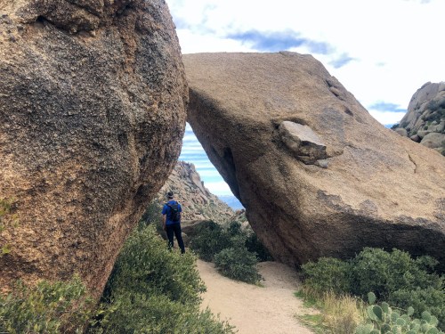 A hiker stands between two large boulders on a rocky trail, surrounded by shrubs and a cloudy sky.