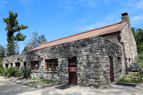 A stone building with a red roof, surrounded by trees and blue sky, featuring large windows and a front door.