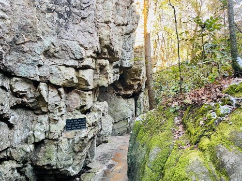 A narrow path between rocky cliffs, with a sign on the left and moss-covered stones, surrounded by trees.