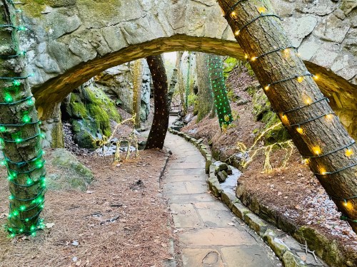 A winding stone path under an archway, flanked by trees wrapped in green and yellow lights, surrounded by mossy rocks.