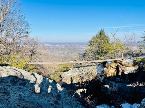 A scenic view from a rocky overlook, showcasing a valley and distant hills under a clear blue sky.