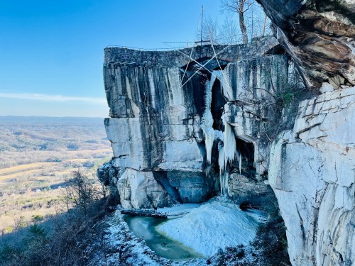 A rocky cliff with icicles hanging down, overlooking a valley with trees and a clear blue sky.