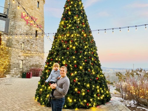 A parent holds a child in front of a large, decorated Christmas tree with lights, set against a scenic backdrop.