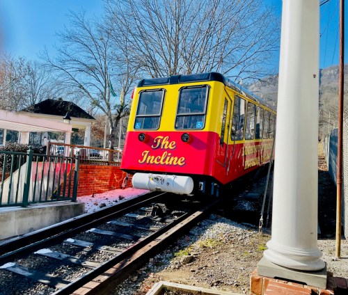 A colorful tram labeled "The Incline" on tracks, surrounded by trees and a building in a sunny setting.