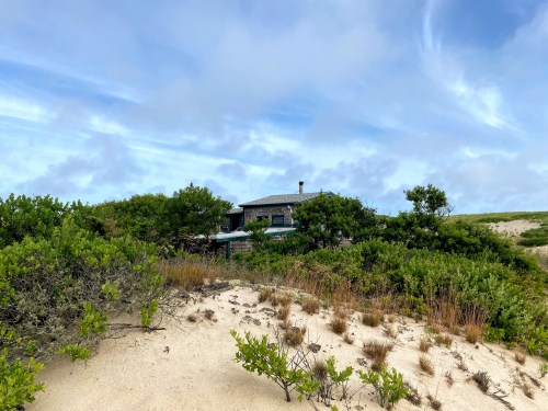 A house nestled among green shrubs and sandy dunes under a cloudy blue sky.