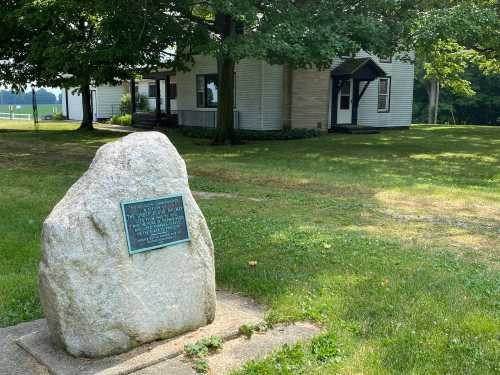 A large rock with a plaque stands in front of a gray house surrounded by trees and grass.