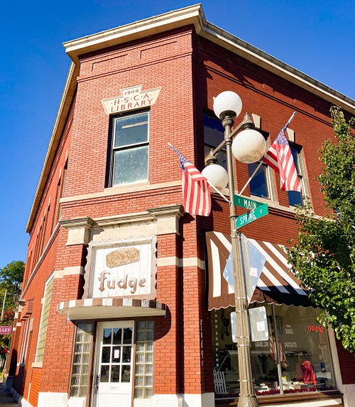 Historic red brick building with a sign for "fudge," American flags, and a corner street sign for Main and Spring.