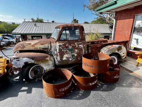 A rusty vintage pickup truck parked beside large, weathered metal rings at a rustic outdoor setting.