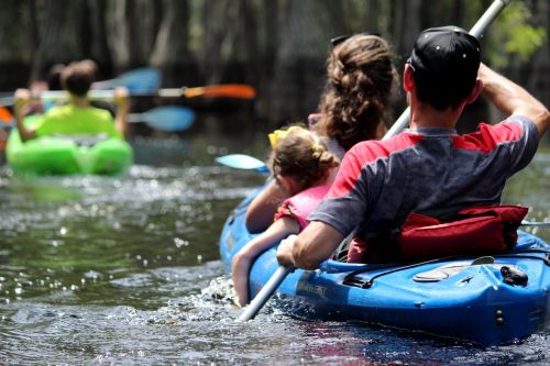 A family kayaking on a calm waterway, surrounded by trees, enjoying a sunny day outdoors.