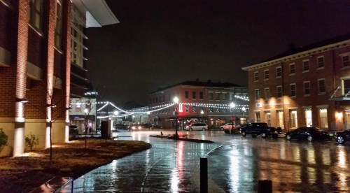 A rainy night scene featuring illuminated buildings and wet streets, with cars parked along the curb.
