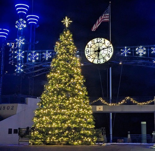 A brightly lit Christmas tree stands at night, with a clock and festive decorations in the background.