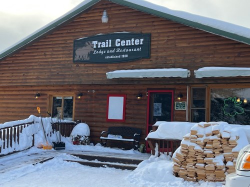A wooden lodge with a sign reading "Trail Center Lodge and Restaurant," surrounded by snow and stacked firewood.