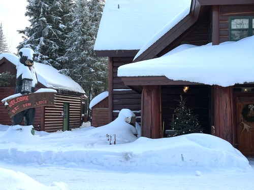 A snowy scene featuring a rustic lodge with a bear statue and a decorated Christmas tree by the entrance.