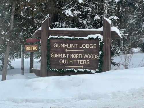 Wooden sign for Gunflint Lodge and Gunflint Northwoods Outfitters, with a bistro sign, surrounded by snow and trees.