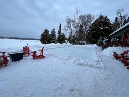 A snowy outdoor scene featuring a frozen ice rink surrounded by red chairs and trees in the background.