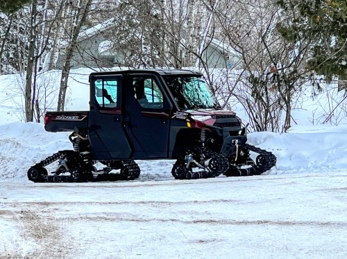 A black all-terrain vehicle with snow tracks parked on a snowy path surrounded by trees.