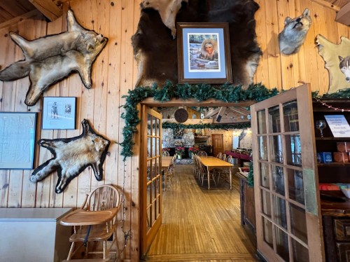 Interior of a rustic lodge with wooden walls, animal hides on display, and a dining area visible through double doors.