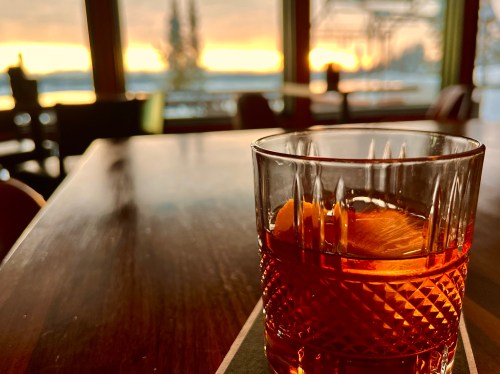 A close-up of a cocktail glass with an orange twist, set on a wooden table with a sunset view in the background.