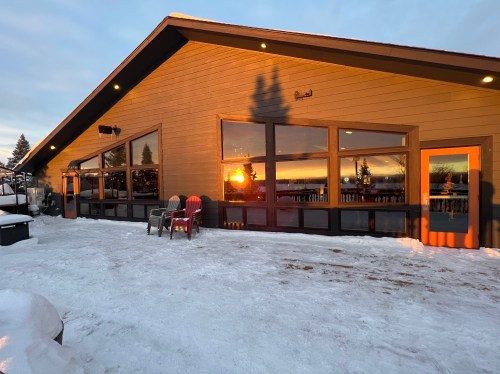 A cozy cabin with large windows reflecting a sunset, surrounded by snow and trees. Adirondack chairs are in front.