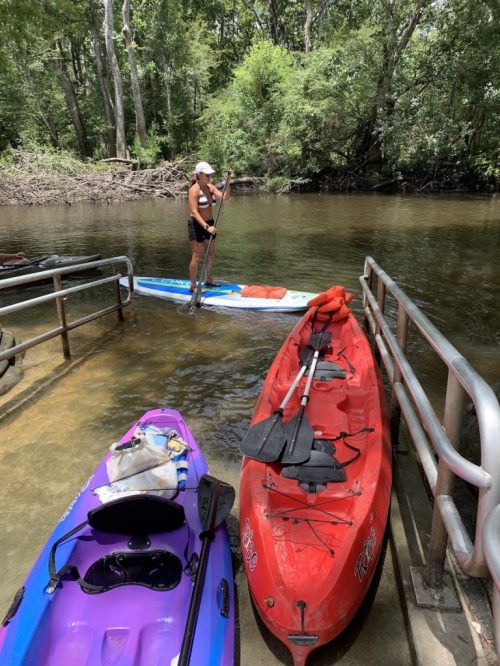 A person stands on a paddleboard near two kayaks by a calm river surrounded by trees.
