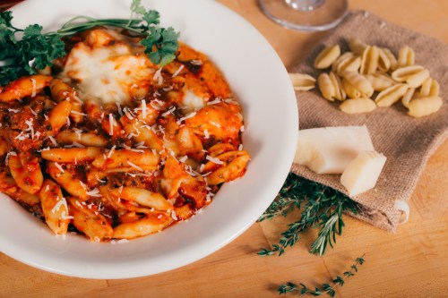 A bowl of pasta with marinara sauce, cheese, and herbs, accompanied by uncooked pasta and cheese on a wooden surface.