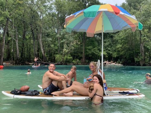 A family enjoys a sunny day on a paddleboard with an umbrella, surrounded by water and trees.