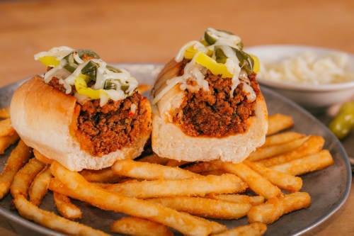 Two loaded hot dog buns filled with seasoned meat, topped with jalapeños and mustard, served with crispy French fries.