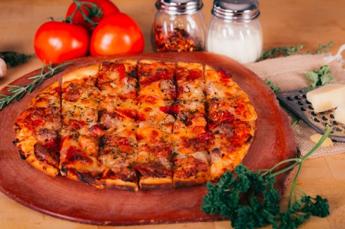 A freshly baked pizza topped with sausage, cheese, and herbs, surrounded by tomatoes and condiments on a wooden board.
