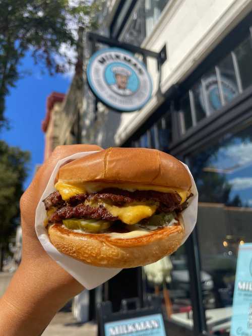 A hand holding a cheeseburger with melted cheese and pickles, in front of a restaurant sign.