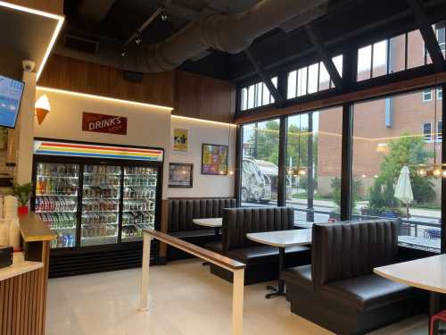 Interior of a modern café with seating booths, a drink fridge, and large windows showing an outdoor view.