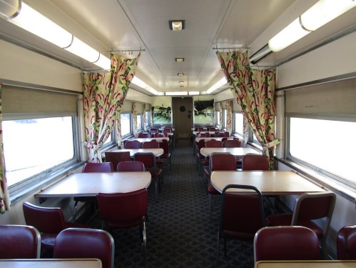 Interior of a vintage train dining car with tables, chairs, and floral curtains, featuring large windows.