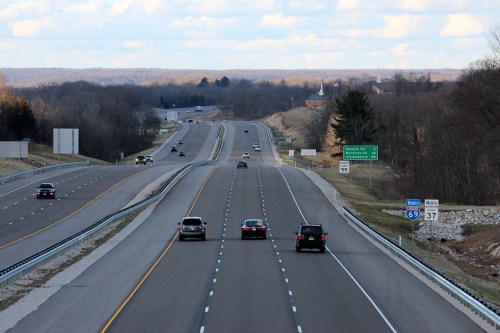 A view of a wide highway with cars traveling, flanked by trees and signs for upcoming exits.