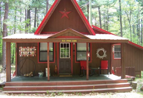 A rustic cabin with a red star, surrounded by trees, featuring a porch and red chairs.