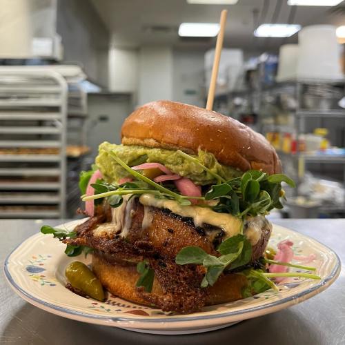 A gourmet burger with grilled meat, guacamole, pickled onions, and microgreens on a toasted bun, served on a plate.