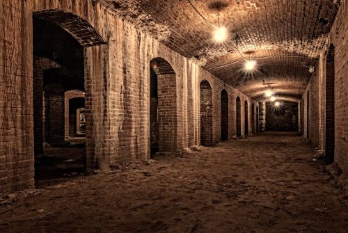 Dimly lit underground corridor with brick arches and exposed light bulbs, featuring a dusty floor and rustic walls.