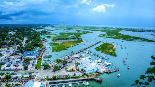 Aerial view of a coastal area with boats, a marina, and lush green marshlands under a cloudy sky.
