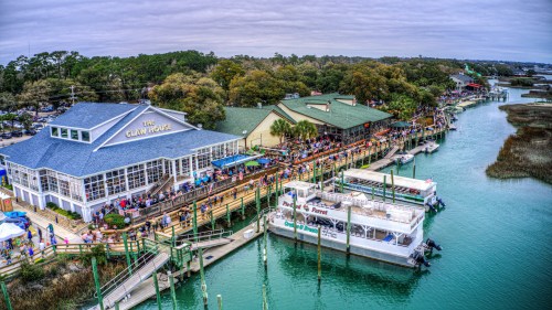 Aerial view of a bustling waterfront restaurant with boats docked, surrounded by trees and a cloudy sky.