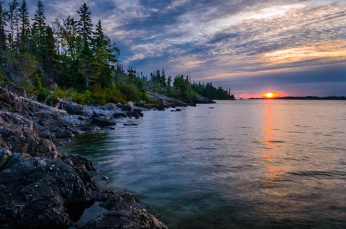 A serene lakeside view at sunset, with rocky shores and trees silhouetted against a colorful sky.
