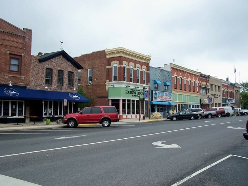A quaint downtown street lined with brick buildings, shops, and parked cars under a cloudy sky.
