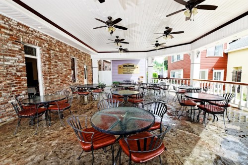 Outdoor dining area with round tables and chairs, featuring a brick wall and ceiling fans, under a covered porch.