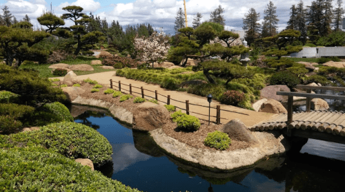 A serene garden with a winding path, a wooden bridge, lush greenery, and a tranquil pond under a partly cloudy sky.