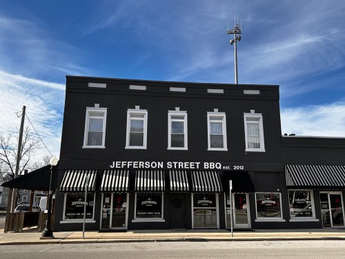 Exterior of Jefferson Street BBQ, a black building with white stripes, established in 2012, under a blue sky.