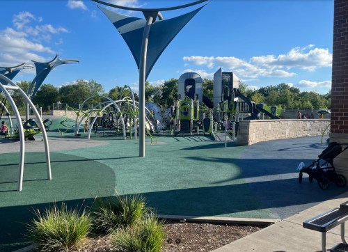 A modern playground with green equipment, shaded areas, and a stroller parked nearby under a clear blue sky.